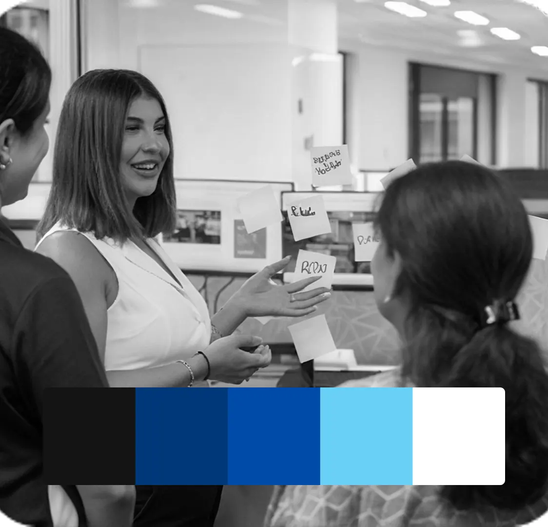A black and white photograph of colleagues talking beside sticky notes on a whiteboard, with a five-colour swatch strip across the bottom.