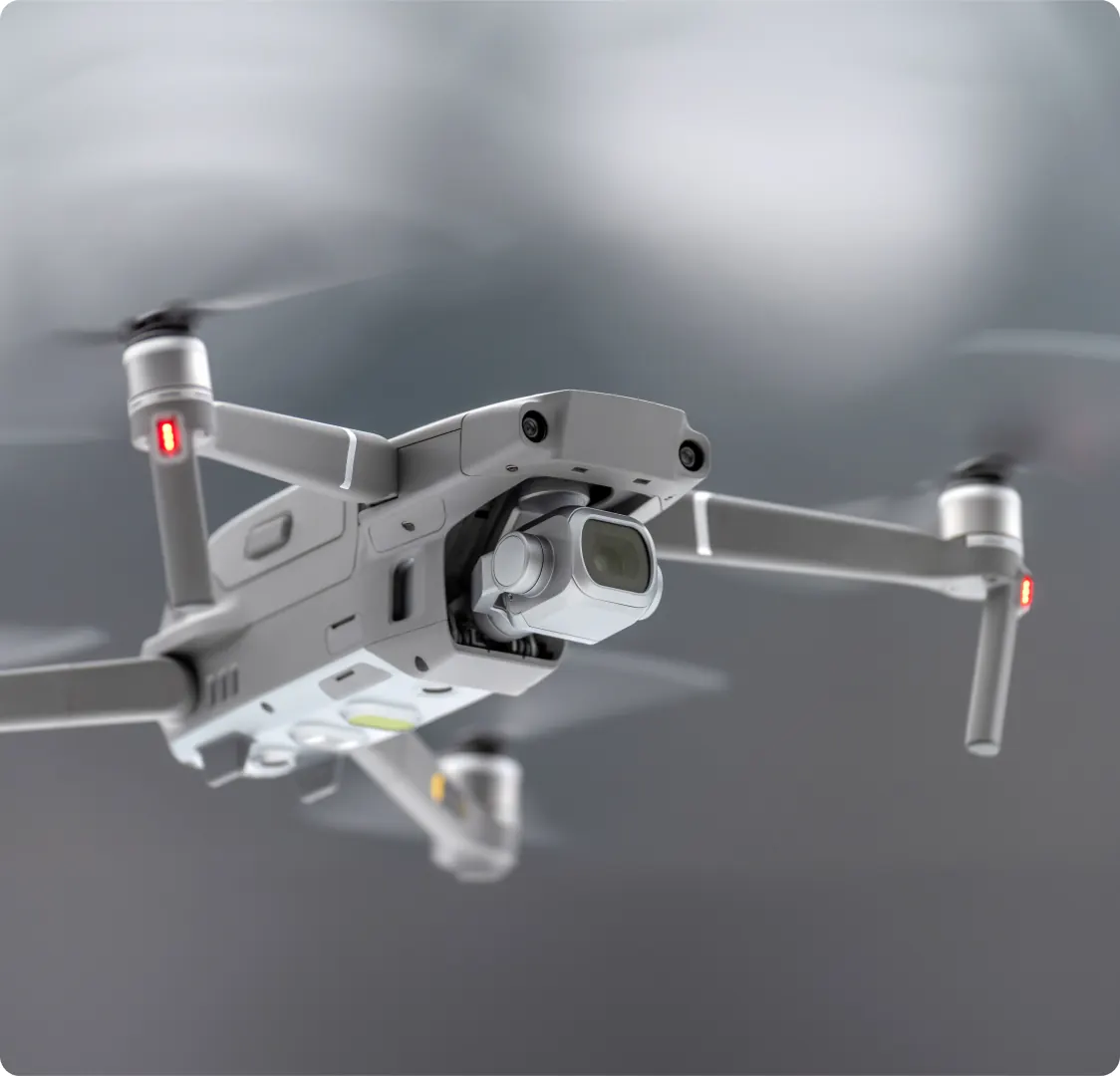 A close photograph of a grey camera drone in flight against a blurred sky.
